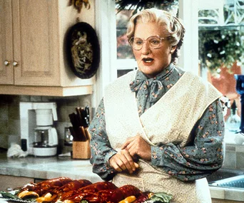 Mrs. Doubtfire cooking in a kitchen, wearing glasses and a floral apron, with lobsters on the counter.