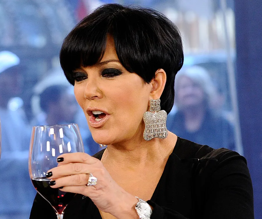 ```plaintext
Person holding a glass of red wine, wearing large diamond earrings and dark attire, talking animatedly.
```