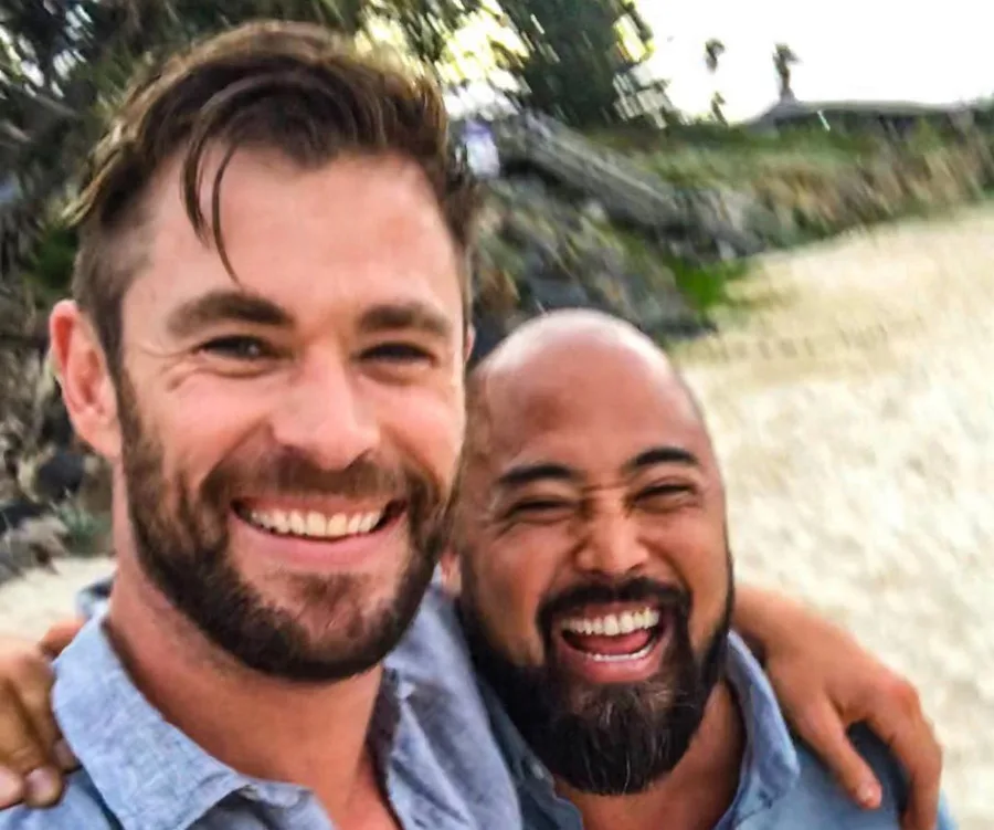 Two men smiling together on a beach, one with a beard and blue shirt, the other with a shaved head and gray shirt.