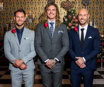 Three men in suits with red roses pinned, standing in elegant room.