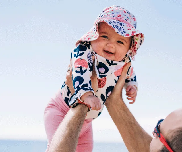 A smiling baby in a sun hat and patterned onesie is held up by an adult with sunglasses against a clear blue sky.