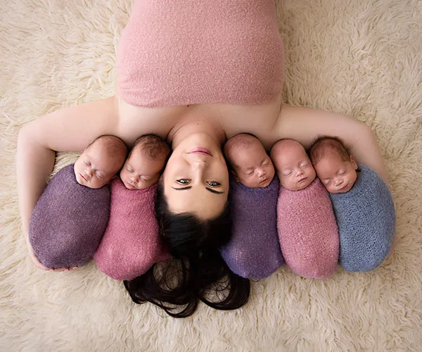Mother lying on carpet with five sleeping babies wrapped in colorful blankets beside her.