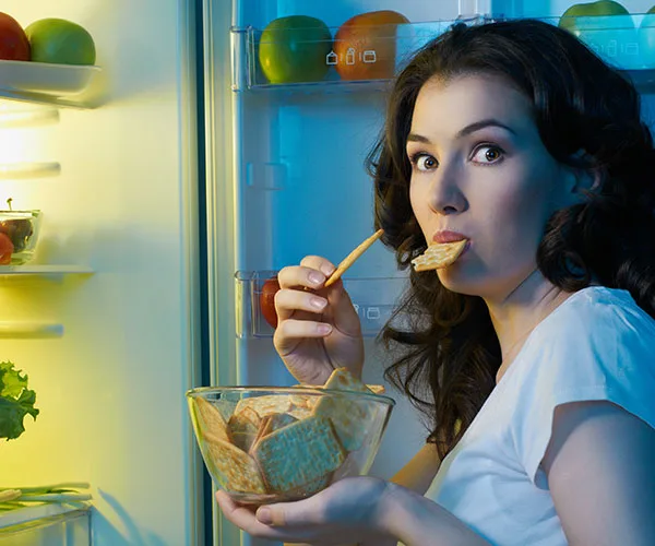 A woman holding a bowl of crackers stands in front of an open fridge, eating one under a cool blue light.