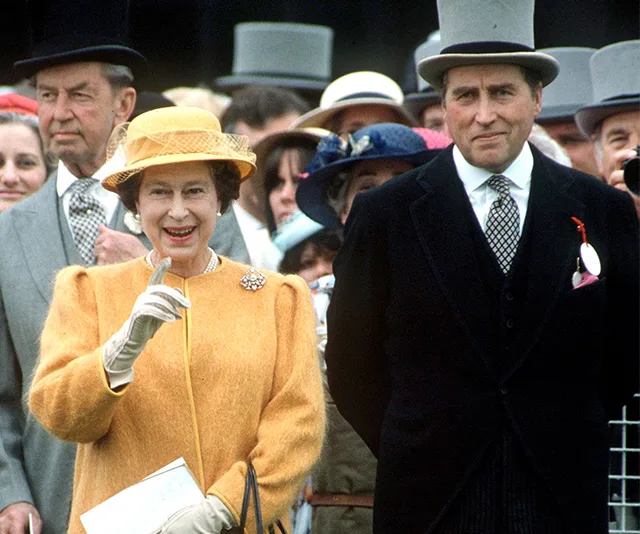 Queen Elizabeth II in a yellow outfit, smiling beside a man in formal attire at an outdoor event with a crowd.