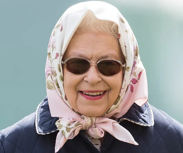 An elderly woman wearing a floral headscarf, sunglasses, and a blue jacket, smiling outdoors.