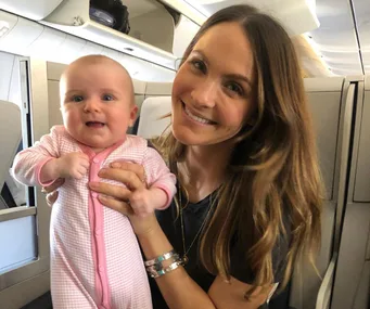 Smiling woman holding a baby dressed in a pink onesie inside an airplane.