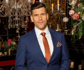 A man in a blue suit with an orange tie stands in a dimly lit, elegantly decorated room.