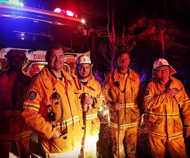 Firefighters in yellow gear stand together at night, lit by emergency vehicle lights, with trees and equipment behind them.