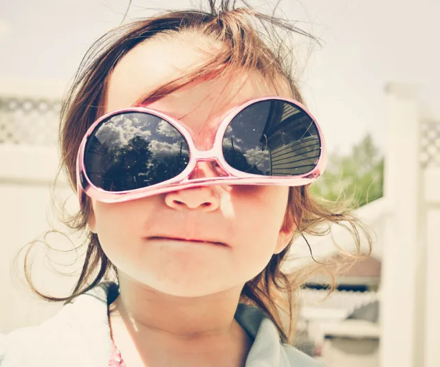 Young child wearing oversized pink sunglasses, smiling outdoors on a sunny day.