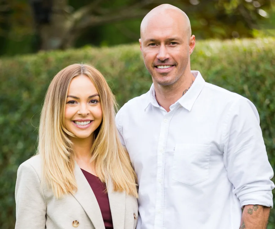 A smiling couple stands together outdoors; woman in a beige blazer and man in a white shirt.