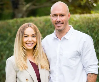 A smiling couple stands together outdoors; woman in a beige blazer and man in a white shirt.