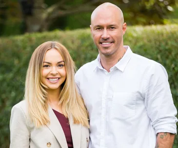 A smiling couple stands together outdoors; woman in a beige blazer and man in a white shirt.