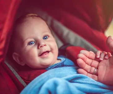 Baby smiling in a red pram, wearing a blue outfit, with an adult's hand nearby.