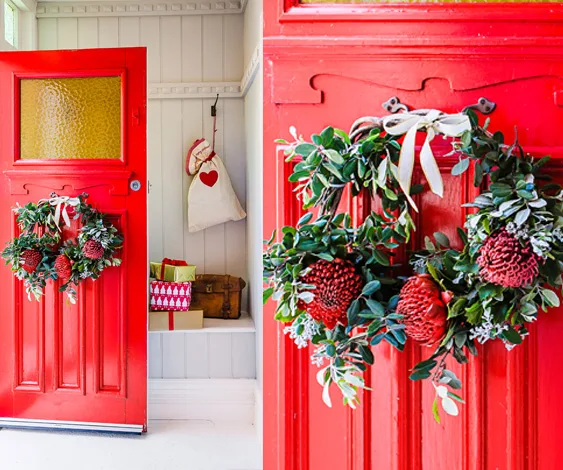 Red door with two fresh flower wreaths featuring greenery and red flowers; festive decorations and gift boxes nearby.