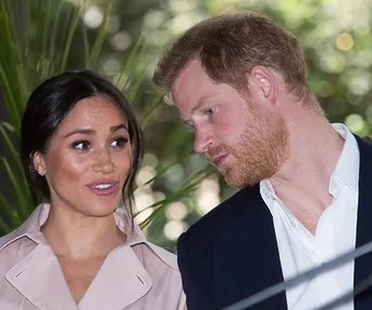 Meghan Markle and Prince Harry at an outdoor event, surrounded by greenery, engaged in conversation.