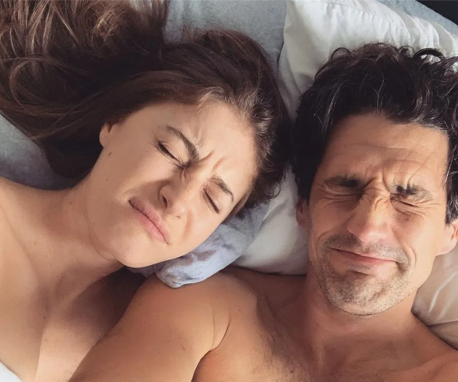 A couple lying in bed with scrunched faces, appearing playful and relaxed.