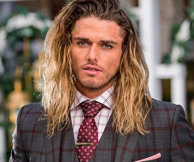 Young man with long blond hair in a checkered suit and patterned tie, standing outdoors.