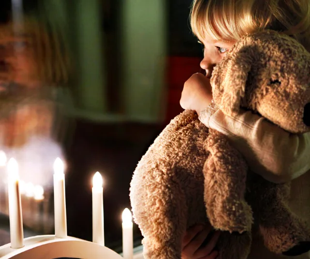 Child hugging teddy bear, gazing out window with lit candles reflected on glass.