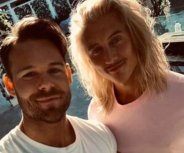Two individuals outdoors smiling for a selfie, one with long blonde hair, the other with short dark hair.