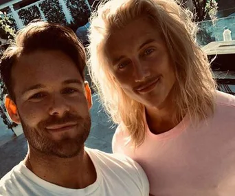 Two individuals outdoors smiling for a selfie, one with long blonde hair, the other with short dark hair.