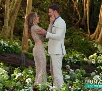 A couple stands facing each other in a garden, surrounded by flowers and trees, both dressed in formal attire.