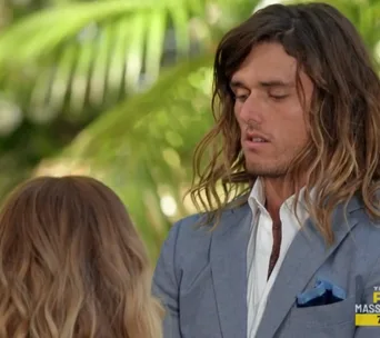 Two people facing each other, one with long hair in a suit, outdoors with greenery in the background.