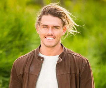 Man with long hair, wearing a brown jacket and white sweater, smiling outdoors with greenery in the background.