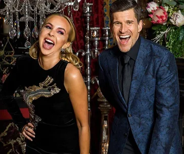 Two people smile and pose in formal attire, standing indoors with decorative elements around them.
