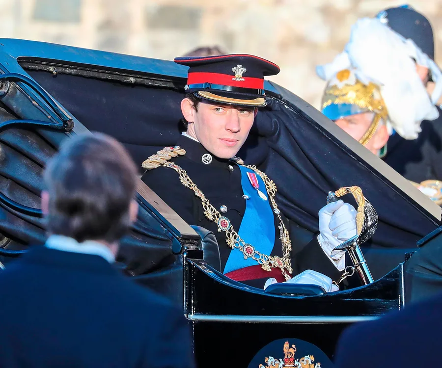 Man in military uniform with red and blue details sits in a carriage, holding a sword, surrounded by onlookers.
