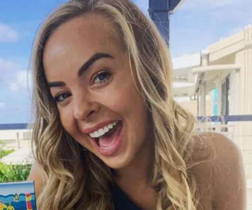 A smiling woman with long blonde hair, enjoying a sunny day outdoors.