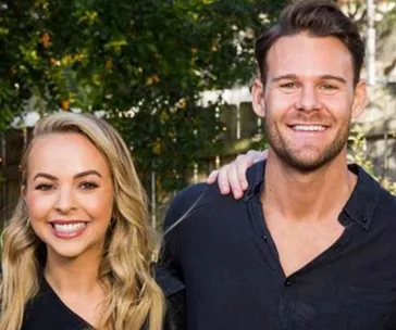 A smiling couple, a blonde woman and a brunette man, stand together outdoors, wearing dark shirts.