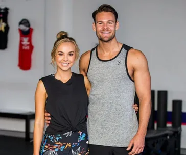 A man and woman smiling and posing in gym clothing inside a workout area.