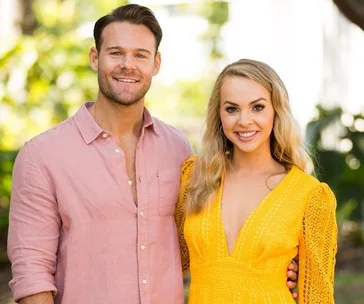 A smiling man in a pink shirt stands with a woman in a yellow dress outdoors.