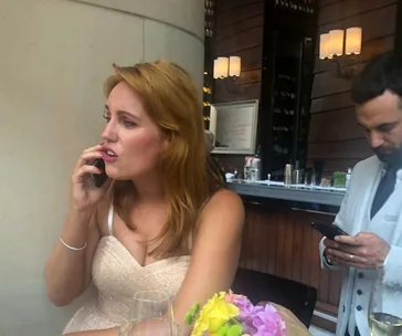 Woman in a dress talks on phone, looking concerned, while a man checks his phone at a bar.