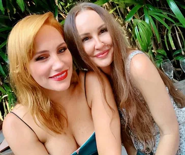 Two women smiling together, with green leafy plants in the background.