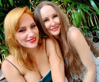 Two women smiling together, with green leafy plants in the background.