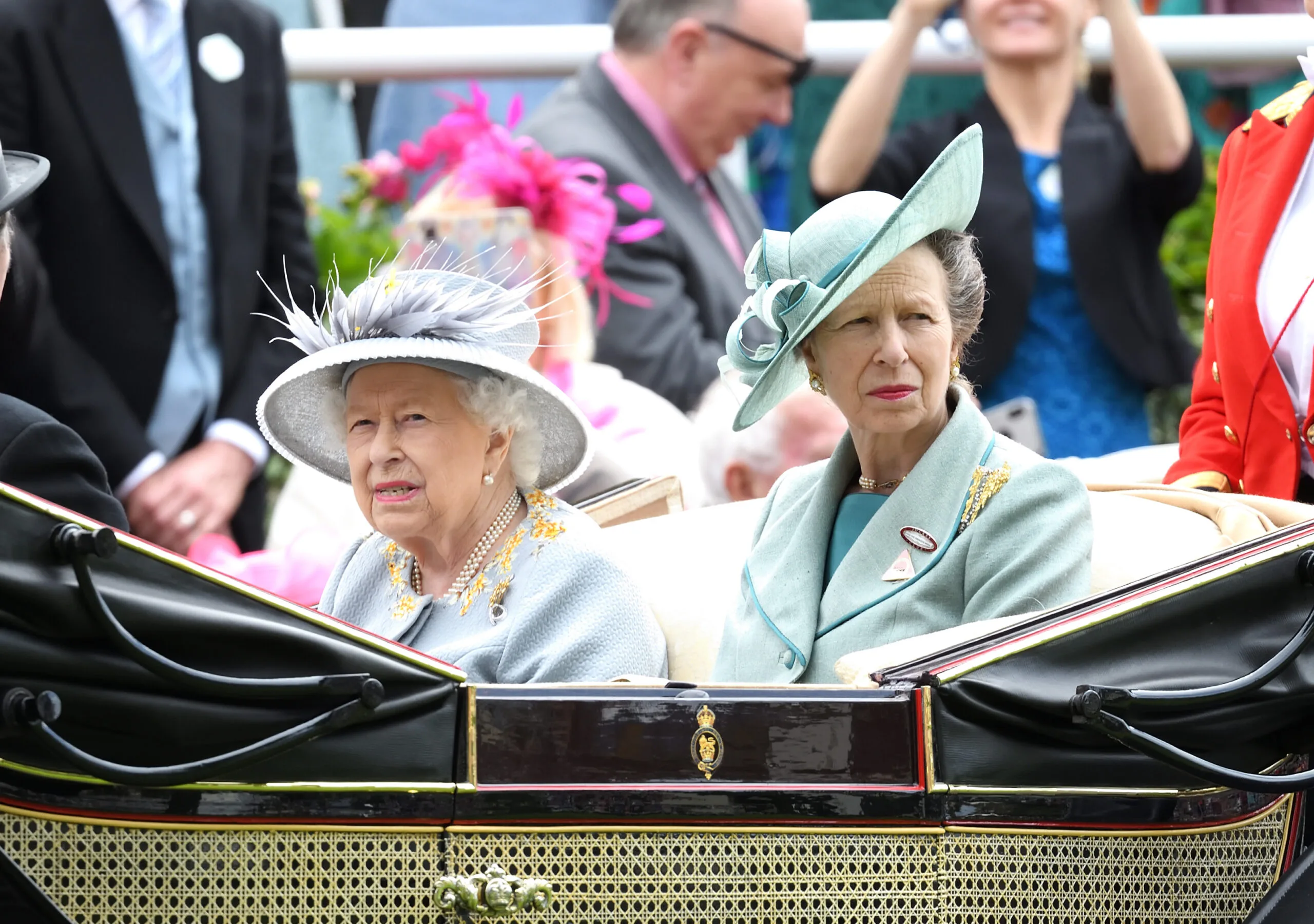 Two women in elegant hats sitting in a carriage at an outdoor event, with people in the background.