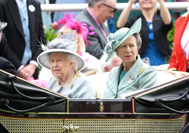 Two women in elegant hats sitting in a carriage at an outdoor event, with people in the background.