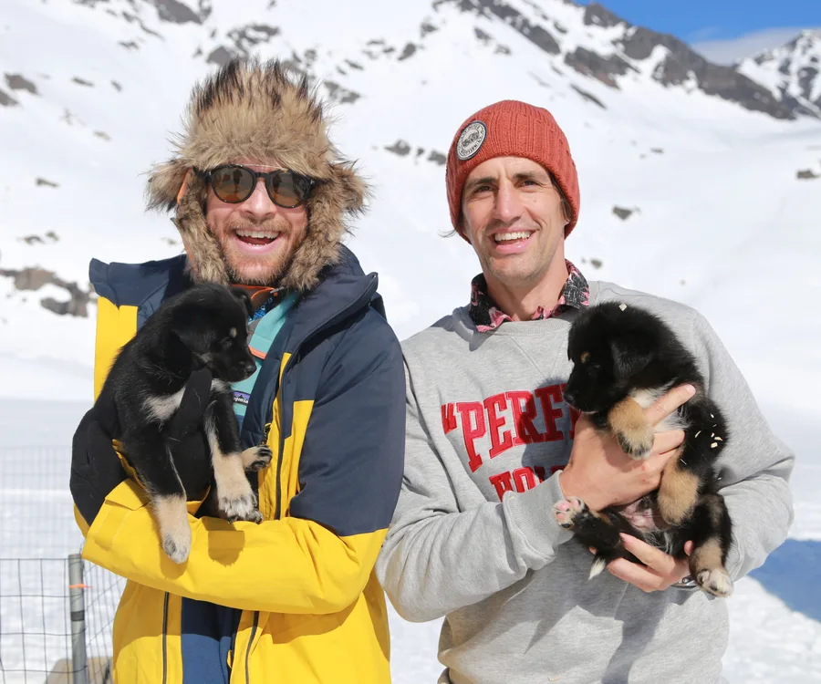 Men in winter clothing holding puppies in a snowy mountain setting, both smiling.