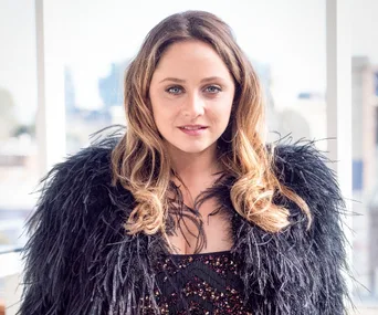 Cece Peters in a feathered black outfit, smiling, with a blurred cityscape in the background.