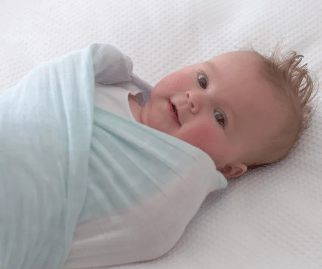 A baby wrapped in a light blue swaddle lying on a white textured blanket, looking up and smiling.