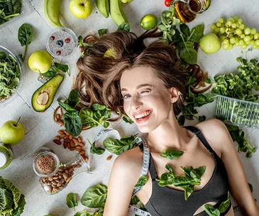 Woman lying on the floor surrounded by fresh fruits, vegetables, nuts, and seeds, smiling and winking.