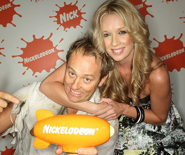 Two people smiling, holding a Nickelodeon blimp award against a backdrop with Nickelodeon logos.