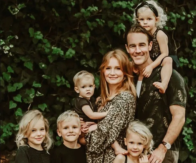 A family of seven, including parents and five children, pose outdoors with greenery in the background.