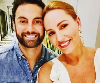 Two people smiling for a selfie indoors, the woman wearing a white dress and hoop earrings, and the man in a denim shirt.