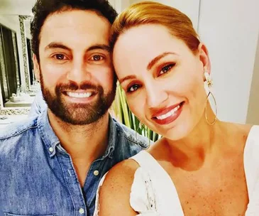 Two people smiling for a selfie indoors, the woman wearing a white dress and hoop earrings, and the man in a denim shirt.