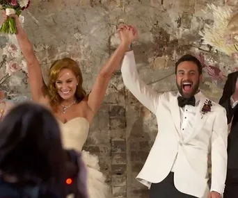 Couple in wedding attire holding hands joyfully, in front of a rustic wall with floral decorations.