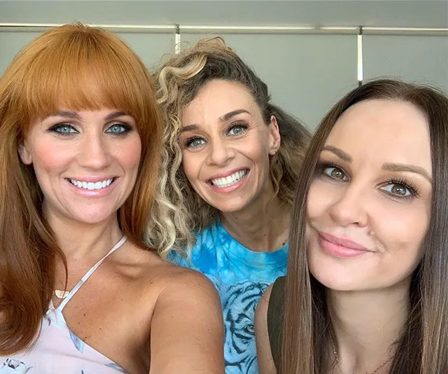 Three women smiling for a selfie, with one wearing a blue shirt and the others in light-colored tops.