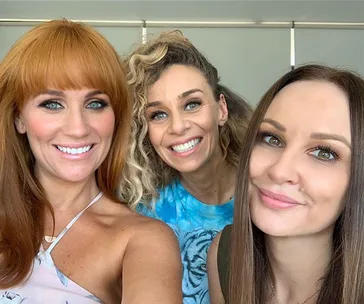 Three women smiling for a selfie, with one wearing a blue shirt and the others in light-colored tops.