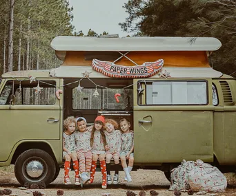 Kids in festive outfits sitting in a vintage van with "Paper Wings" sign, surrounded by trees and decorations.